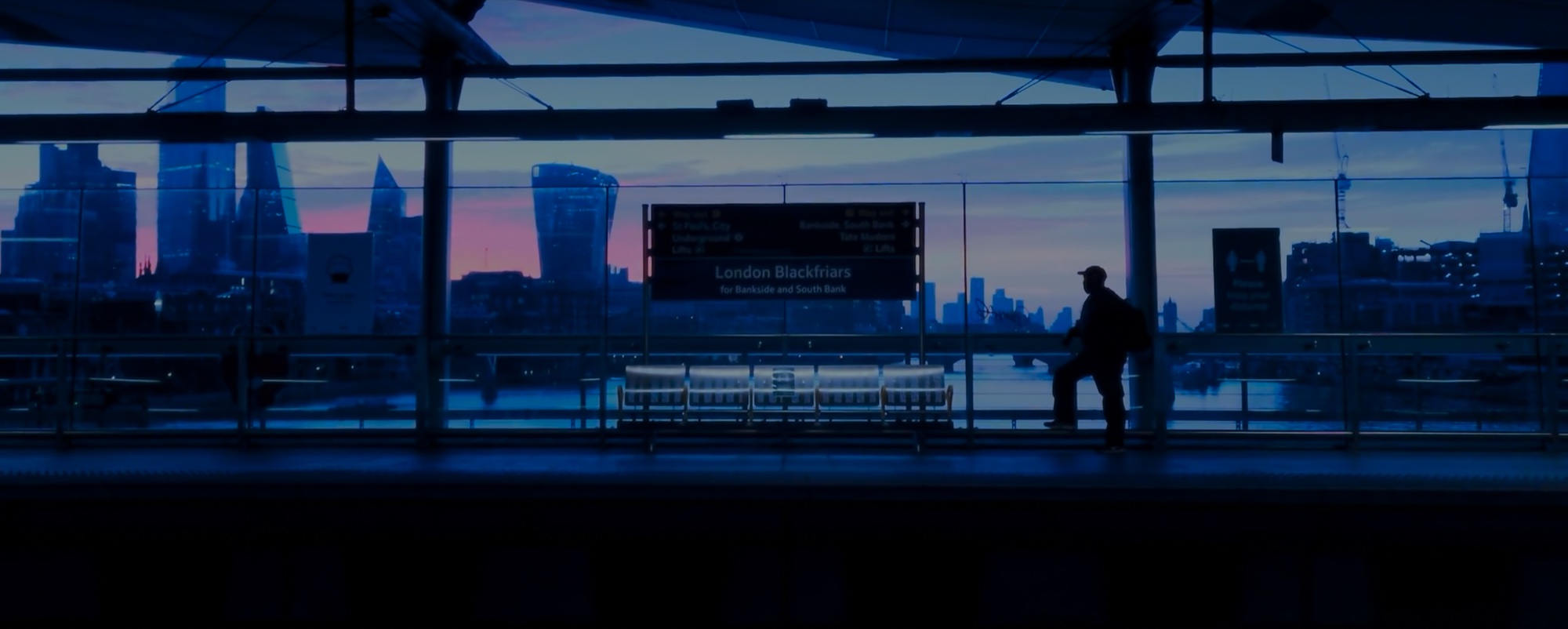 London skyline at dusk through the girders of a station roof, a lone figure on the platform