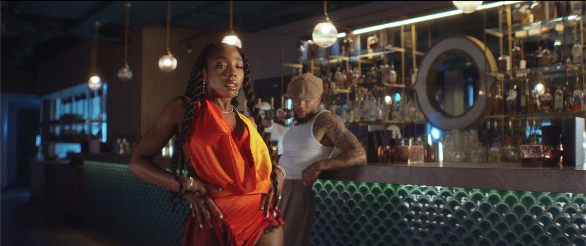 Nissi in an orange dress at a green-tiled bar, warm practicals behind