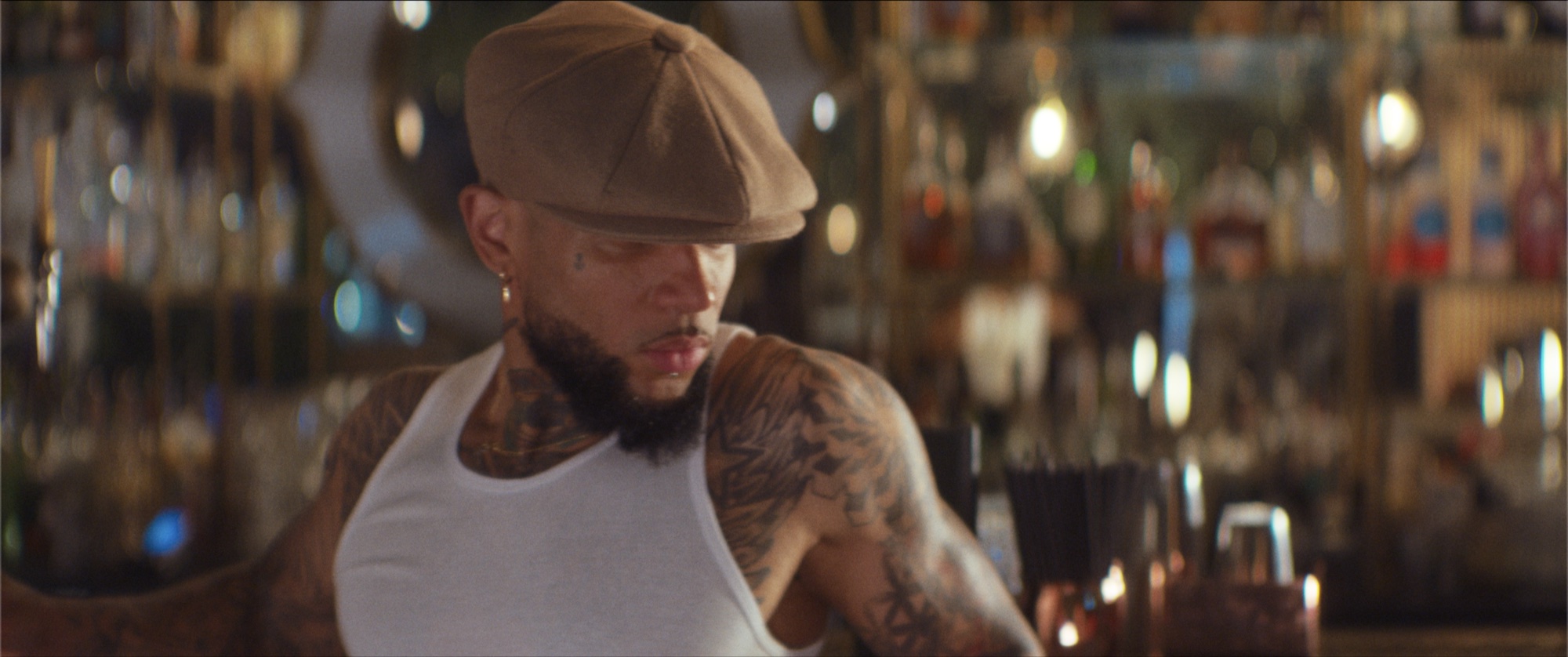 Man in a flat cap and white vest leaning across the bar